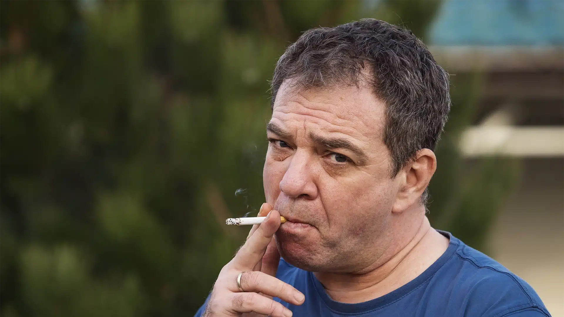 hypnosis, hypnotherapy, health A man in a blue shirt is smoking a cigarette outdoors, with greenery in the background, indulging in smoking as a false relaxant.