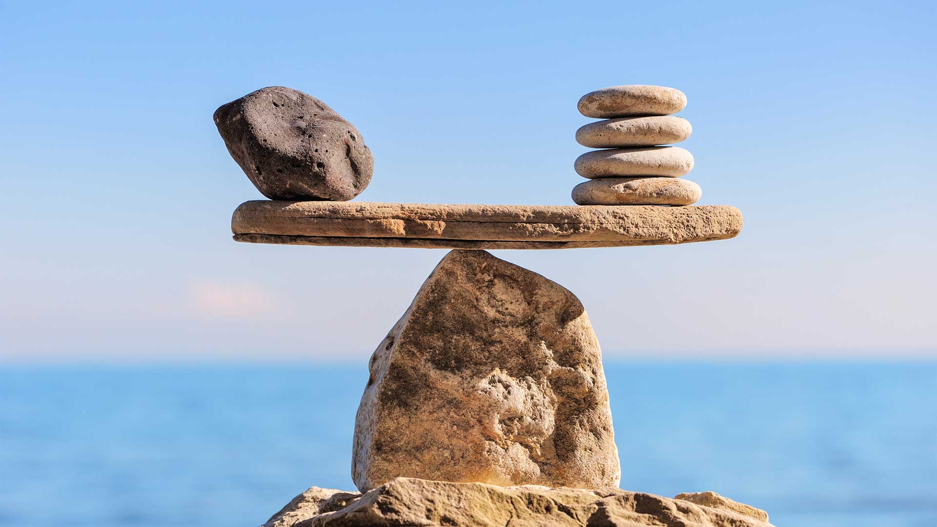 Hypnosis Tune-Up. A large dark stone and a stack of smaller white stones balance precariously on opposite ends of a flat rock, all supported by a larger rock with the sea and sky in the background.