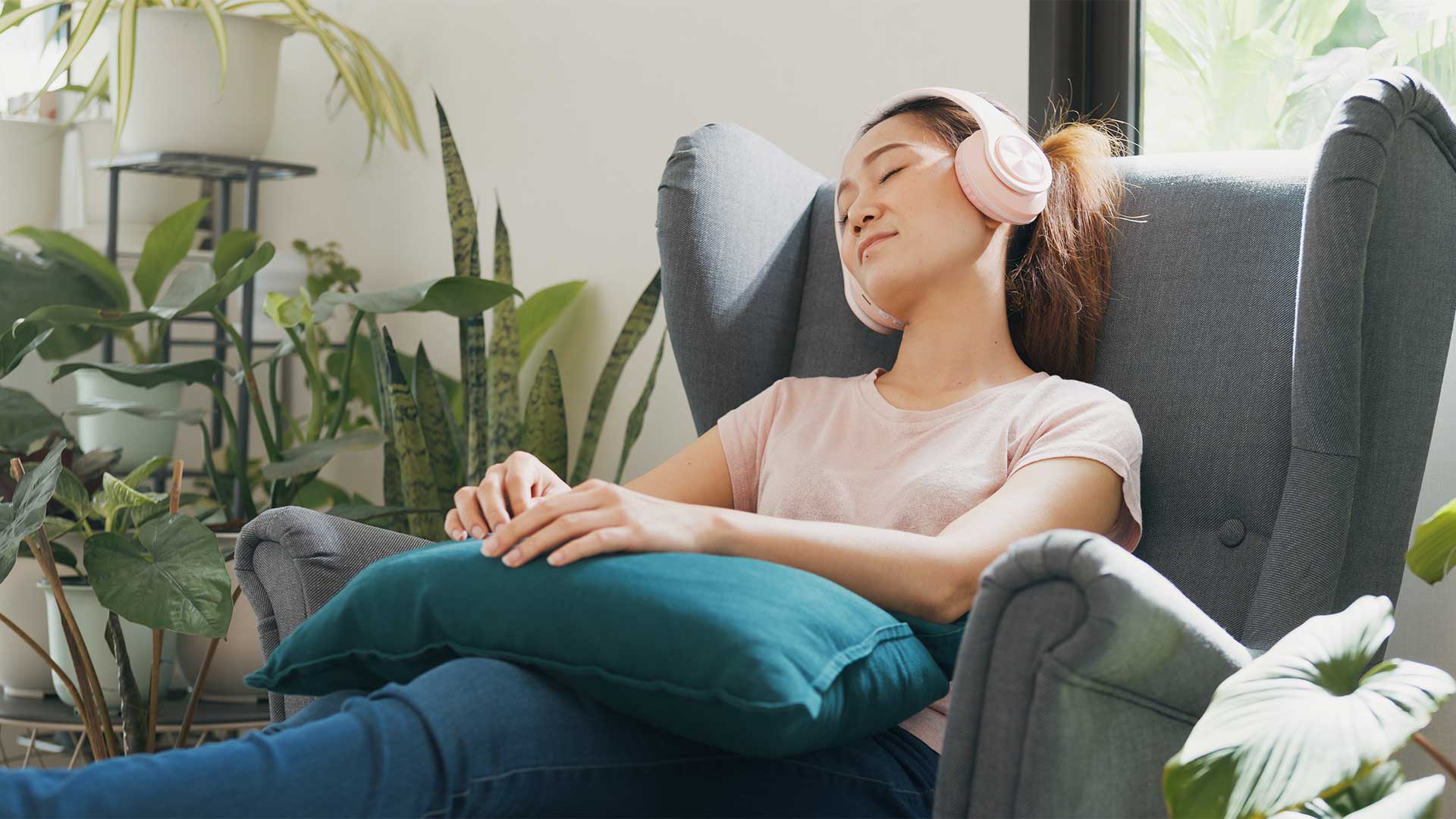 Self-hypnosis, hypnotherapy, health A woman wearing headphones rests with her eyes closed in a cushioned chair, surrounded by houseplants, holding a teal pillow.