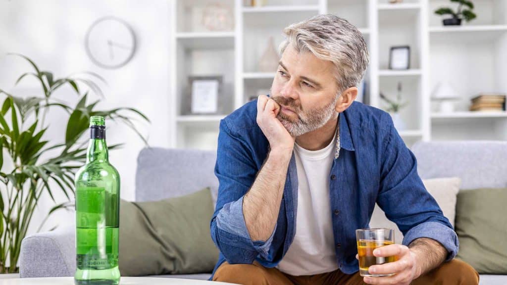 Alcohol Addiction Recovery Process. A man sits on a couch, holding a glass of whiskey, contemplating the journey of alcohol recovery. An open green bottle rests on the table before him as he reflects thoughtfully on his progress.