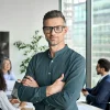 A man wearing glasses and a green shirt stands confidently with arms crossed in an office setting, where his three colleagues are working in the background, possibly discussing the newest approaches from "Hypnosis to be More Assertive.