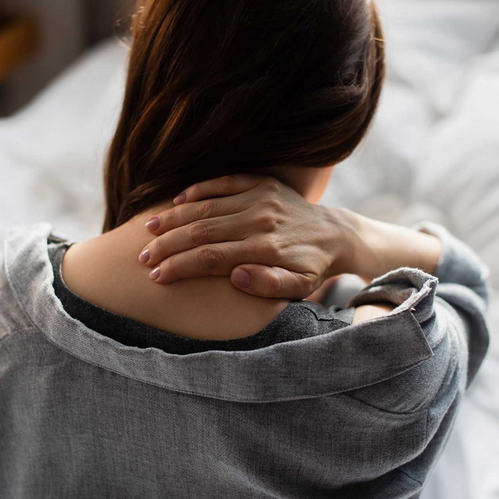 A person with long hair holds the back of their neck with one hand, wearing a grey garment, perhaps seeking relief reminiscent of a calming self-hypnosis download.