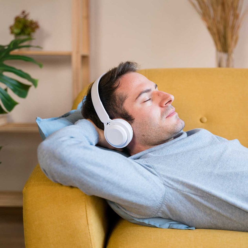 A man wearing white headphones is lying on a yellow couch with his eyes closed and hands behind his head, seemingly immersed in a self-hypnosis download.