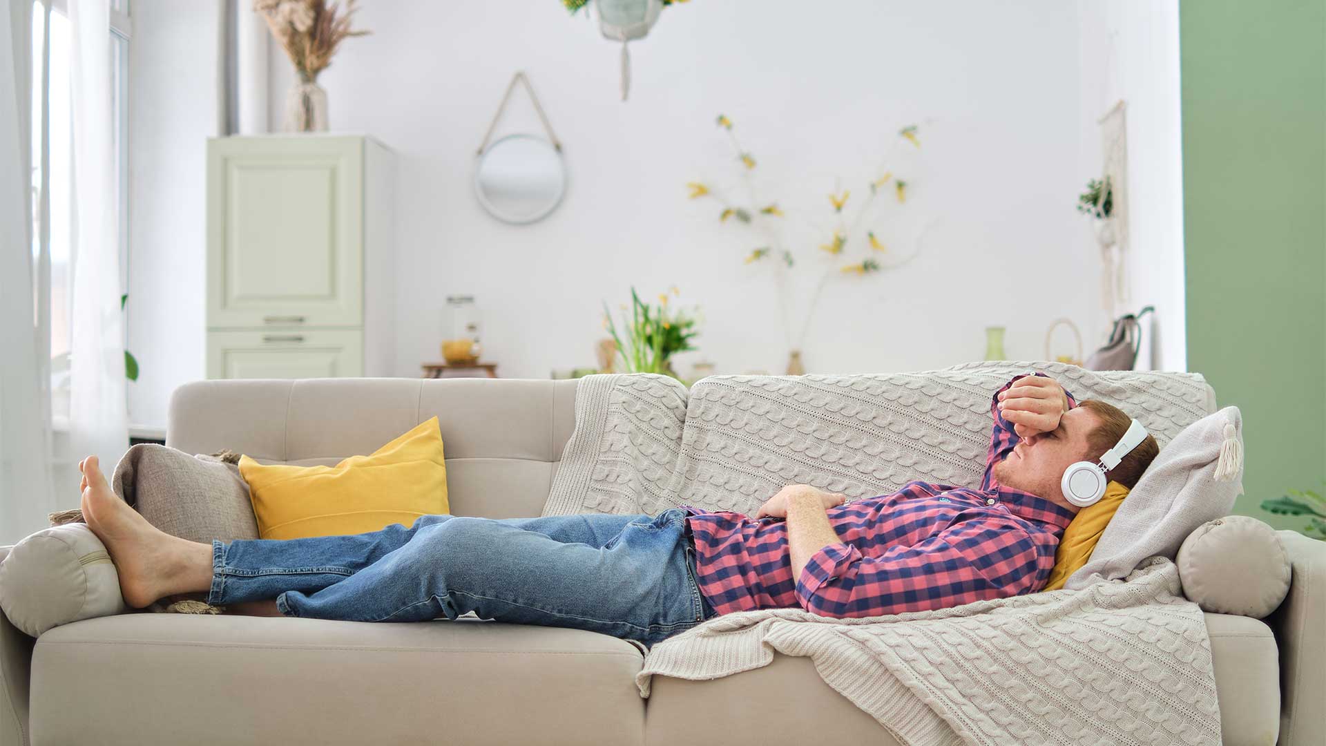 A person wearing headphones and a plaid shirt lies on a beige couch with their feet outstretched and an arm resting over their face, possibly engaging in self-hypnosis.