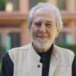 An Older Man With A White Beard And Mustache, Resembling Bruce Lipton, Stands Smiling Outdoors In A Black Shirt And Beige Vest, With A Building And Lush Greenery In The Background.