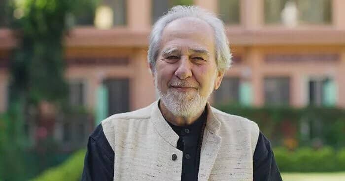 An Older Man With A White Beard And Mustache, Resembling Bruce Lipton, Stands Smiling Outdoors In A Black Shirt And Beige Vest, With A Building And Lush Greenery In The Background.