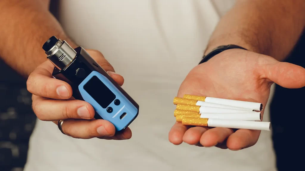 A Man Engaging In Vaping Holds An Electronic Cigarette Alongside A Pack Of Cigarettes.