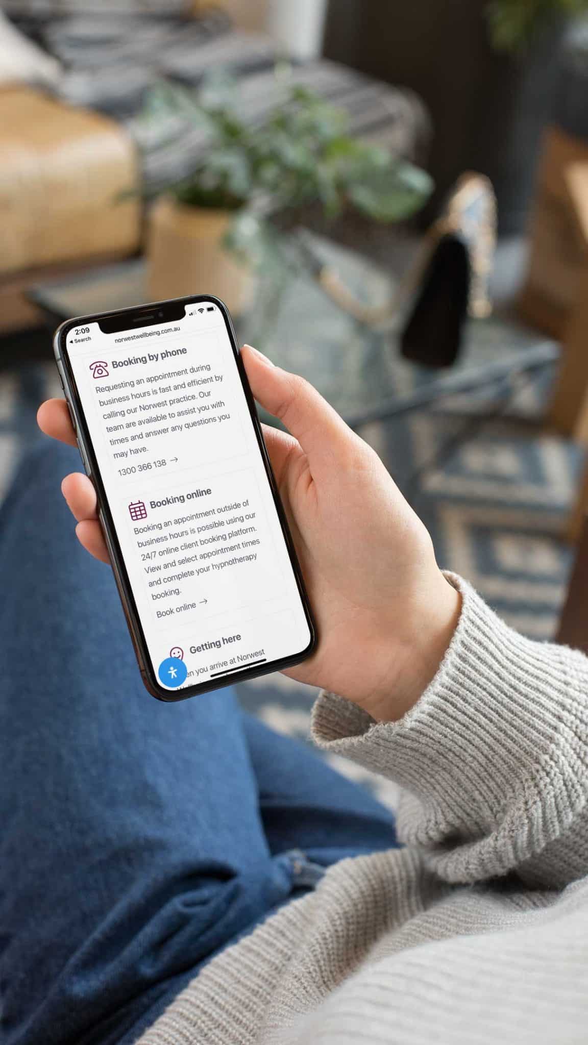 Person Holding A Smartphone Displaying A Webpage With Booking And Location Information For Clinical Appointments. The Individual Is Seated, Wearing A Light-Colored Sweater And Blue Jeans.