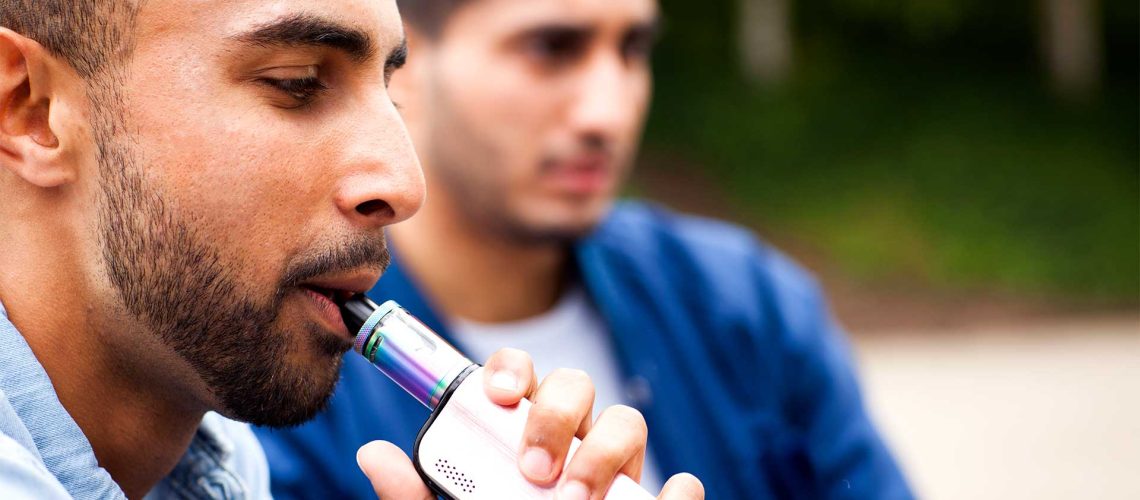 two men sitting next to each other inhaling a vape.